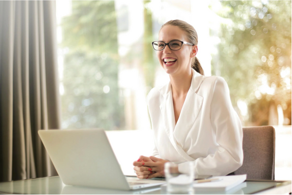 A smartly-dressed, smiling woman sitting at a desk with a laptop in a professional setting